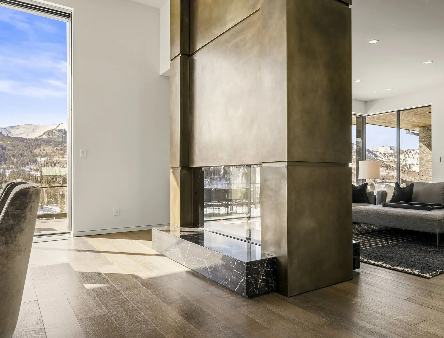 Two-storey patinated bronze fireplace on a black marble hearth, snow-covered ridgeline through the glass