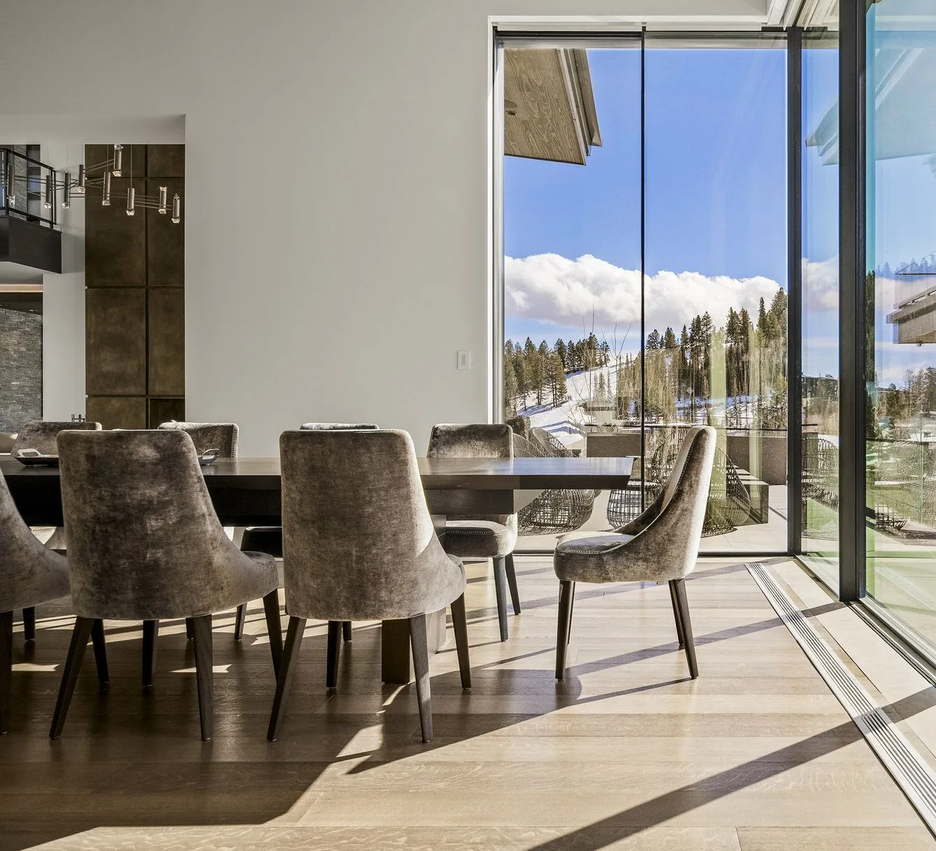 Dining table and velvet chairs beside a glass wall onto snow-covered pines