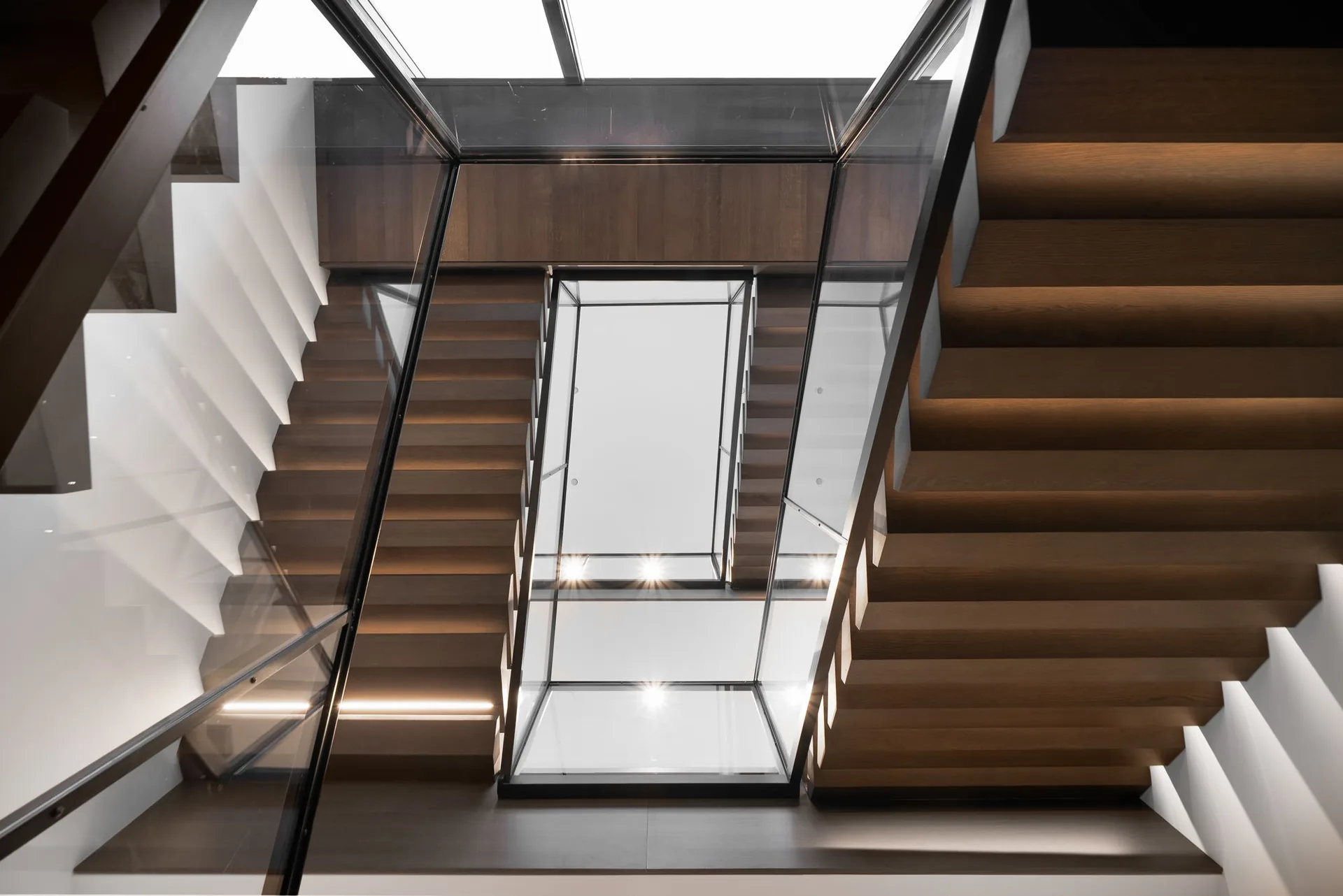 Stairwell seen from above, flights of timber treads flanking the glass elevator shaft